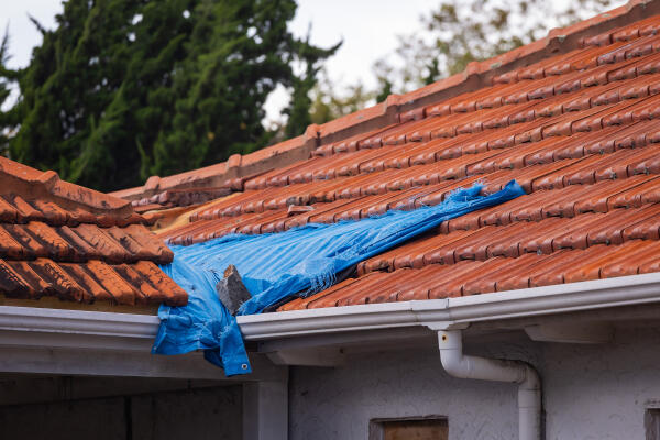 A blue tarpaulin on top of a damaged roof, showing loose roof tiles.