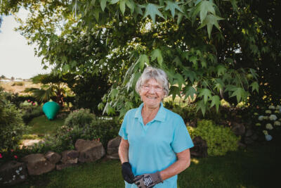 An older woman standing and smiling in her yard, with trees and plants in the background.