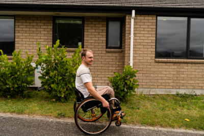 A young man in a wheelchair in front of his brick house.
