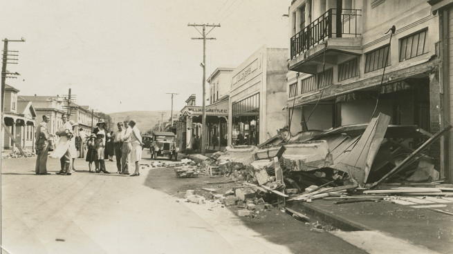 A black and white photo of a street in Napier after the earthquake, with fallen buildings on roads, and a crowd of people standing.