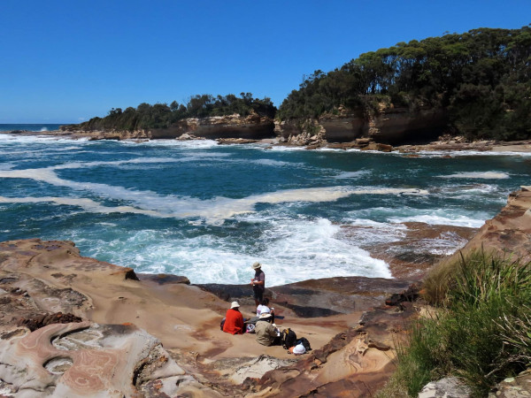 three researchers sit on rock formations next to the sea