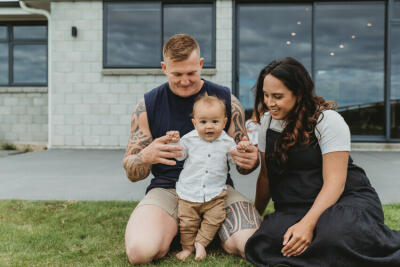 A young man and woman hold their baby, smiling and infront of their home.