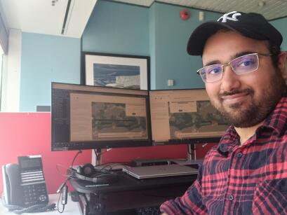 A man smiles at the camera sitting infront of two computer screens.
